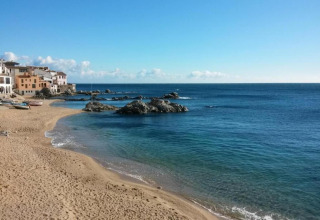 Sandy beach with clear blue water, rocks, and colorful houses beside a bright blue sky and sunshine.