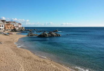 Plage de sable avec eau bleue limpide, rochers et maisons colorées sous un ciel bleu éclatant.