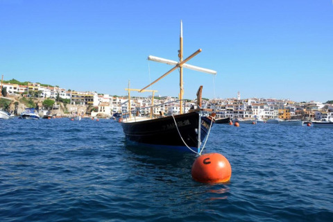 Un bateau traditionnel amarré à une bouée orange flotte sur la mer, avec une ville côtière en arrière-plan.
