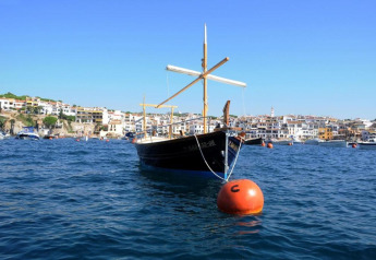 Un bateau traditionnel amarré à une bouée orange flotte sur la mer, avec une ville côtière en arrière-plan.