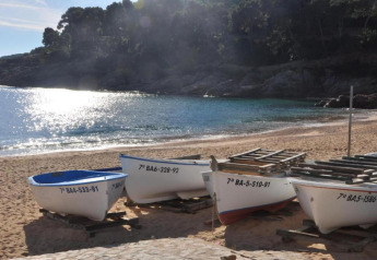 Petits bateaux alignés sur une plage de sable près d’une baie calme, reflet solaire sur l’eau bleue.