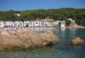 Vue d’une plage avec rochers, eau claire, vacanciers et maisons blanches devant une colline boisée.