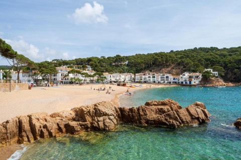 Plage ensoleillée avec eaux claires, rochers et maisons blanches dans un village côtier verdoyant.