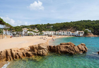 Plage ensoleillée avec eaux claires, rochers et maisons blanches dans un village côtier verdoyant.