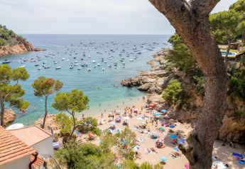 Vue sur une plage animée et mer bleue avec de nombreux bateaux au Resort «Begur 3» en Espagne, entourée d’arbres.