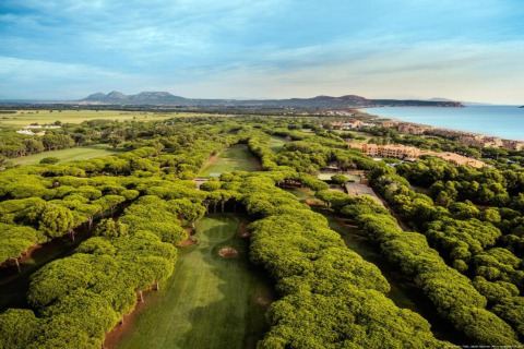 Vue aérienne d’un parcours de golf verdoyant en bord de mer, entouré d'arbres, de montagnes et de bâtiments.