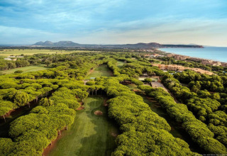 Luchtfoto van een groene golfbaan aan zee, omgeven door bomen, met bergen en gebouwen op de achtergrond.