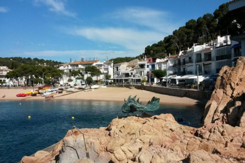 Vue d’un village côtier, maisons blanches, bateaux sur la plage et statue de dragon sur les rochers.