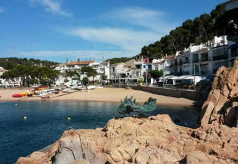 Vue d’un village côtier, maisons blanches, bateaux sur la plage et statue de dragon sur les rochers.