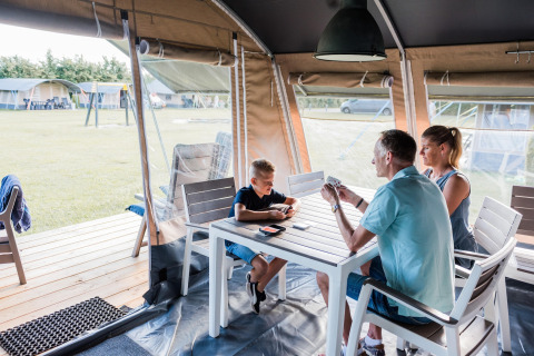 Familia jugando a las cartas en una mesa blanca dentro de una tienda safari en un camping soleado.