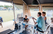 Familia jugando a las cartas en una mesa blanca dentro de una tienda safari en un camping soleado.