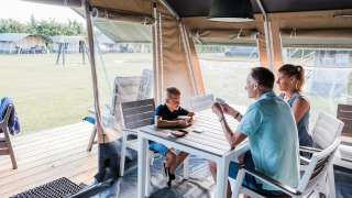 Gezin speelt kaarten aan een witte tafel in een safaritent op een zonnige camping met grasveld.