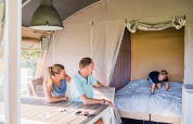 Een familie geniet in een safaritent; ouders zitten aan tafel, terwijl een jongen op het bed speelt.