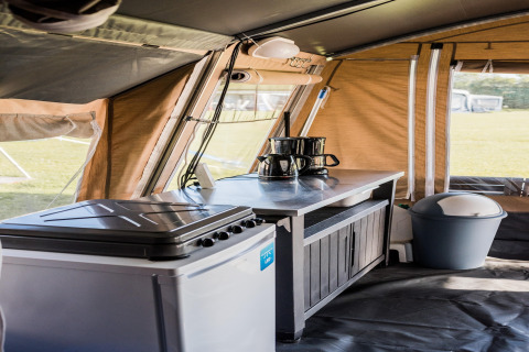 Interior de una tienda safari con zona de cocina pequeña, estufa, cafetera y cubo de basura en un camping.
