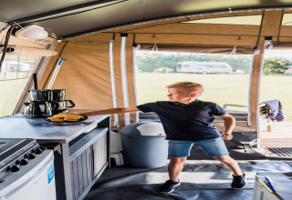 Niño en una tienda safari alcanzando galletas en la mesa, con caravana y camping visible al fondo.