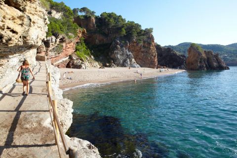 Kvinde går på sti langs klippen med udsigt over stranden og det klare blå vand ved Sa Riera i Spanien.