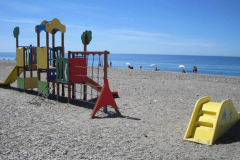 Speeltuin op een kiezelstrand met uitzicht op zee, mensen en parasols op de achtergrond.