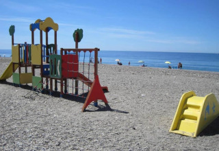 Speeltuin op een kiezelstrand met uitzicht op zee, mensen en parasols op de achtergrond.