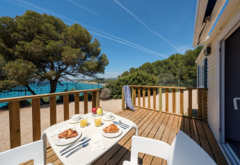 Terrasse en glamping avec petit-déjeuner, croissants, jus, vue sur la mer et arbres en plein soleil.
