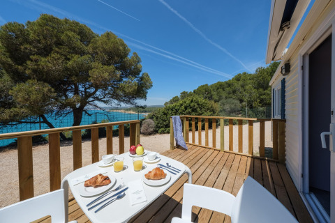Glamping terras met ontbijt, croissants, fruitsap, zicht op zee en bomen onder een blauwe hemel.
