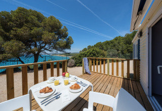 Træterrasse ved glamping, morgenmad med croissanter og juice, udsigt til hav og træer i solen.