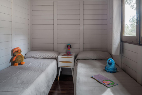 Twee bedden in een gezellige houten bungalow met knuffels, speelgoed en een open boek bij het raam.