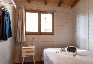 Houten bungalowkamer met bed, raam, nachtkastje en hangende jurken in Torre de la Mora, Spanje.