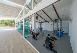 Heldere fitnesszaal met moderne toestellen bij glampingverblijf, grote ramen met zicht op de natuur.