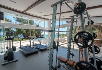 Salle de sport moderne avec tapis de course, haltères et grandes fenêtres donnant sur la mer et les arbres.