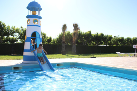 Niños deslizándose por un tobogán con forma de faro que termina en la piscina de un alojamiento turístico.