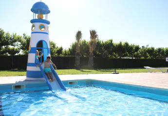 Niños deslizándose por un tobogán con forma de faro que termina en la piscina de un alojamiento turístico.