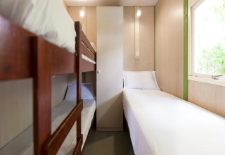 Lodgekamer met houten stapelbed, eenpersoonsbed, witte lakens en groot raam met daglicht.