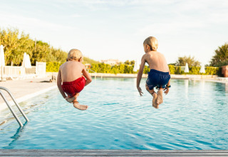 Twee kinderen in rode en blauwe zwembroek springen in het buitenzwembad bij een lodge op een zonnige dag.