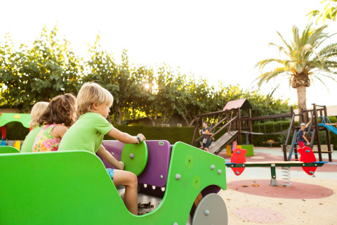Kinderen spelen op een speeltuin bij een lodge, omgeven door palmbomen en speeltoestellen in de zon.