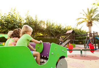 Niños jugando en un parque infantil en una posada, rodeados de palmeras y juegos bajo el sol.