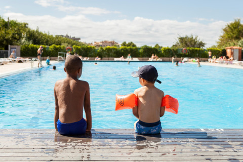 To børn sidder på kanten af en pool i solen ved glamping, det ene barn har badevinger på.