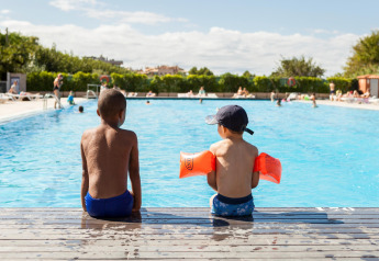 Dos niños están sentados al borde de una piscina en un glamping, uno lleva flotadores naranjas.