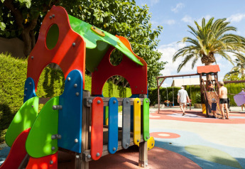 Kleurrijke speeltuin bij een glamping-accommodatie met speeltoestel, palmbomen en mensen op de achtergrond.