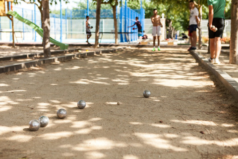 Folk spiller petanque på en solrig grusbane ved et glampingsted med legeplads og tennisbaner i baggrunden.