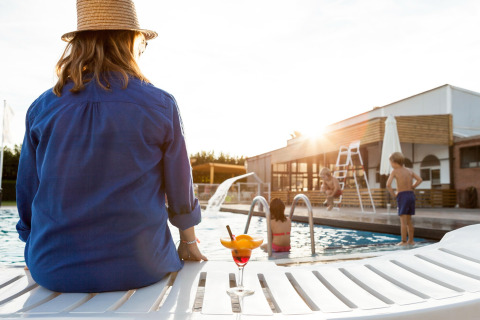 Mujer con sombrero se relaja junto a la piscina de una cabaña mientras los niños juegan al atardecer.