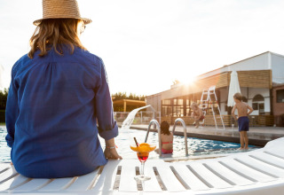 Mujer con sombrero se relaja junto a la piscina de una cabaña mientras los niños juegan al atardecer.
