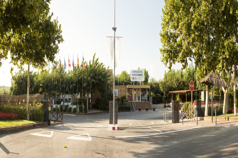 Entrada de Camping Cambrils Playa 1 BUNGALOW OASIS en España con árboles altos, banderas y ambiente soleado.