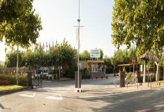 Entrada de Camping Cambrils Playa 1 BUNGALOW OASIS en España con árboles altos, banderas y ambiente soleado.