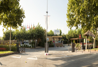 Entrada de Camping Cambrils Playa 1 BUNGALOW OASIS en España con árboles altos, banderas y ambiente soleado.