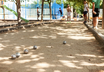 Personas juegan petanca en una cancha de arena bajo sombra de árboles, bolas metálicas dispersas.