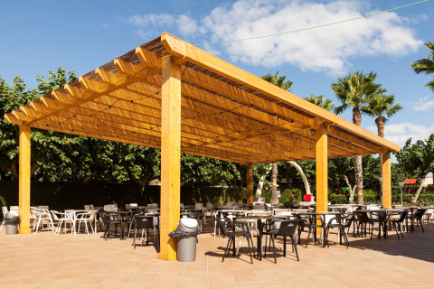 Zona de comedor al aire libre bajo una gran pérgola de madera en un glamping, rodeada de palmeras y árboles.