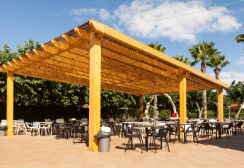 Zona de comedor al aire libre bajo una gran pérgola de madera en un glamping, rodeada de palmeras y árboles.
