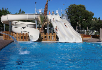 Niños deslizándose por toboganes de agua hacia una piscina en una cabaña, rodeados de árboles y cielo azul.