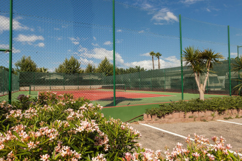 Tennisbaan met groen hekwerk, bloeiende struiken en palmbomen bij Camping Cambrils Playa 2 BUNGALOW PREMIUM in Spanje.