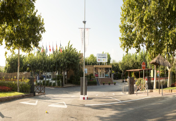 Entrada de un lodge con banderas, árboles y recepción, día soleado y ambiente exterior relajado.
